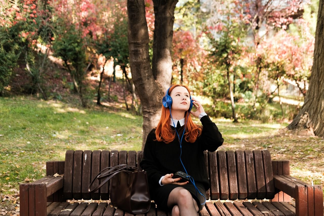 Young woman enjoying music on headphones, seated on a bench in a colorful autumn park.