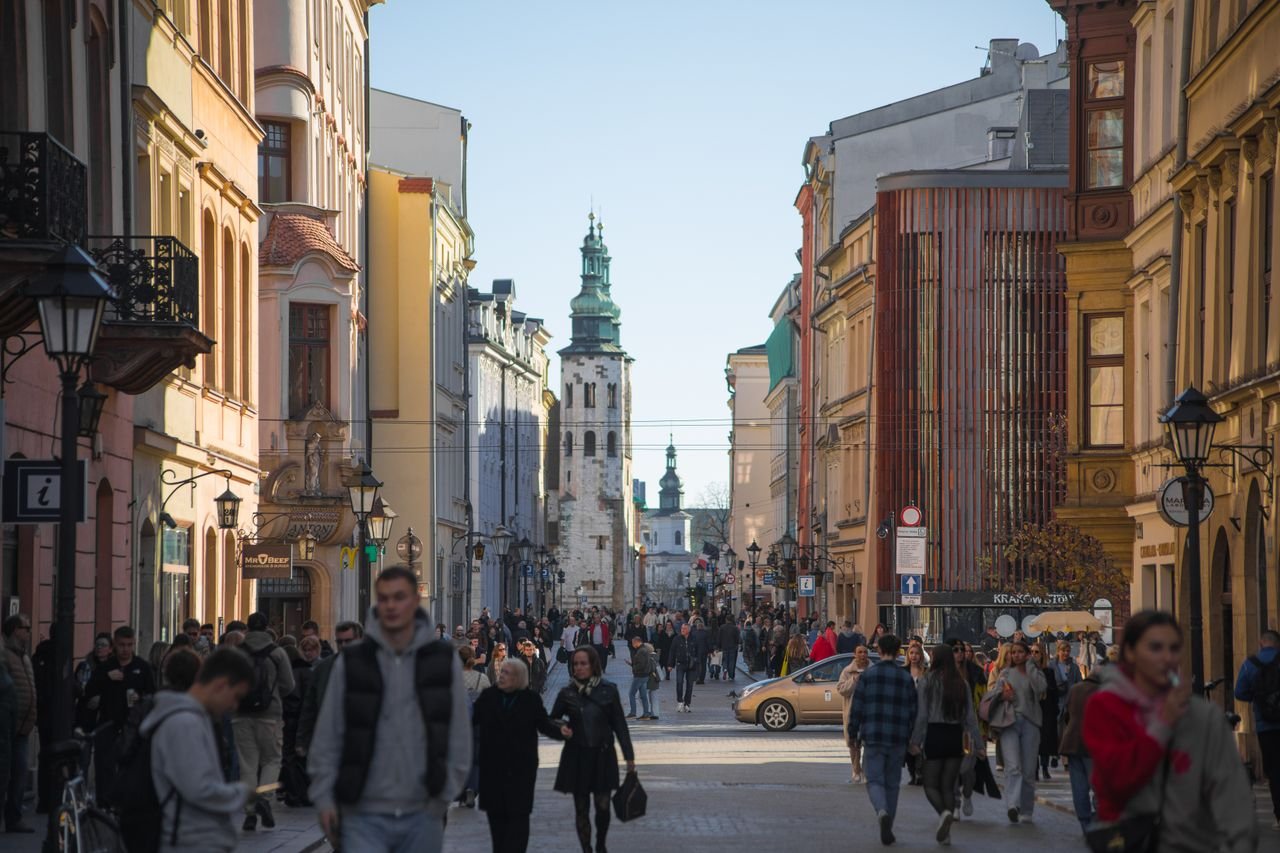 Bustling street with people in a historic city, featuring beautiful architecture and a central tower.